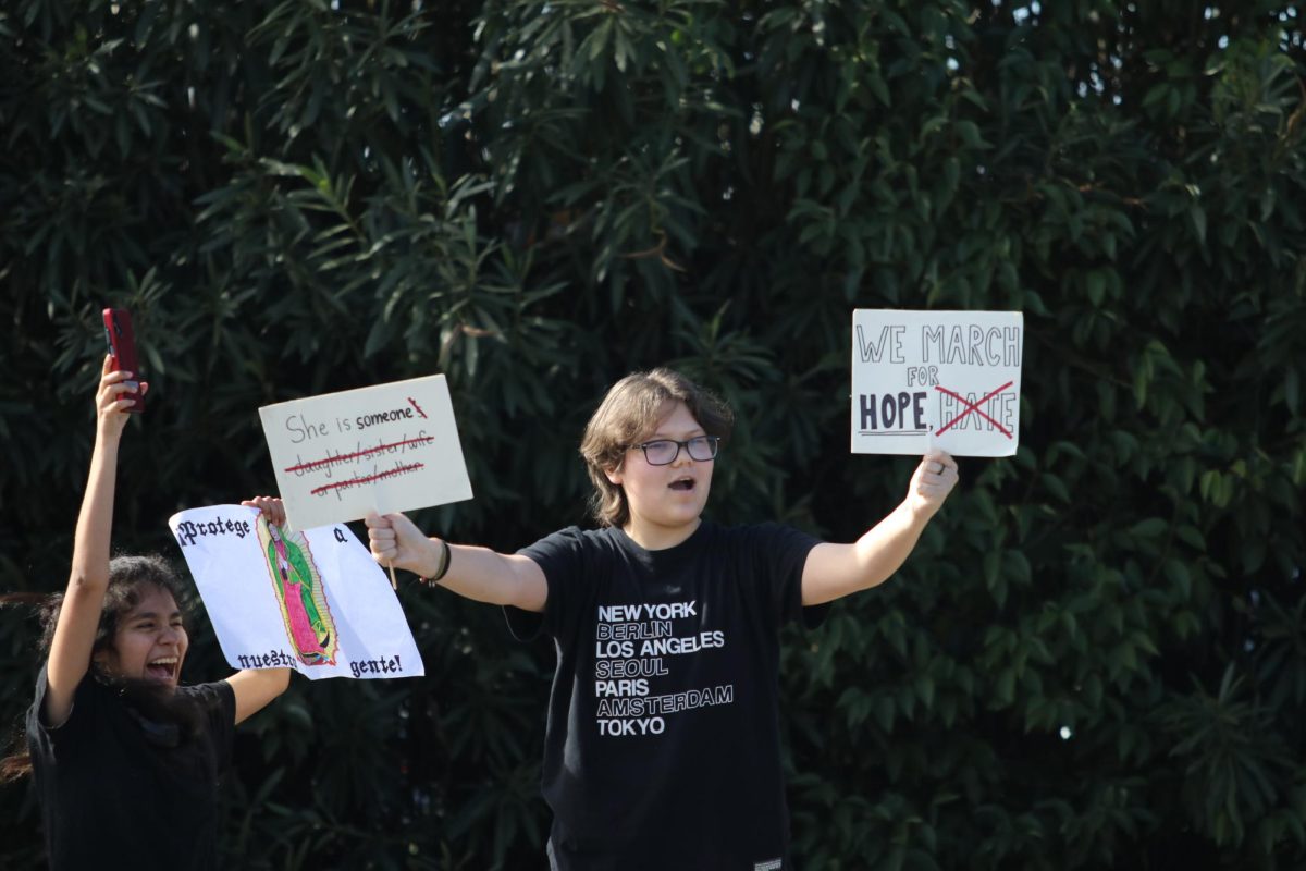 9th grader Adrian Fowler holds up two signs advocating for hope and humanity, while 11th grader Evan Murillo stands beside him with a sign reading “Protect Our People” in Spanish.