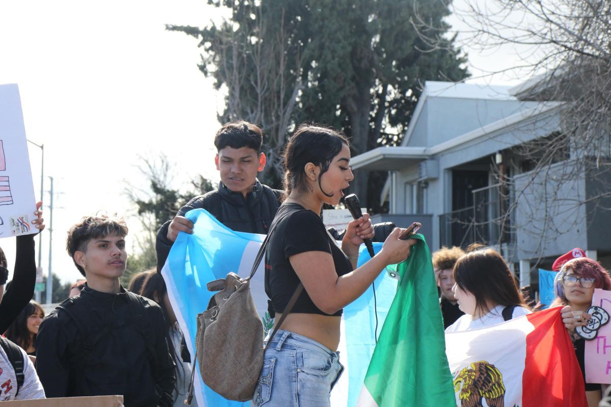 11th grader Amalinalli Peréz stands on a bench, speaking into a microphone. "Thank you for coming hand in hand to put our differences aside and do a peaceful protest for our people to show that we are a united nation."