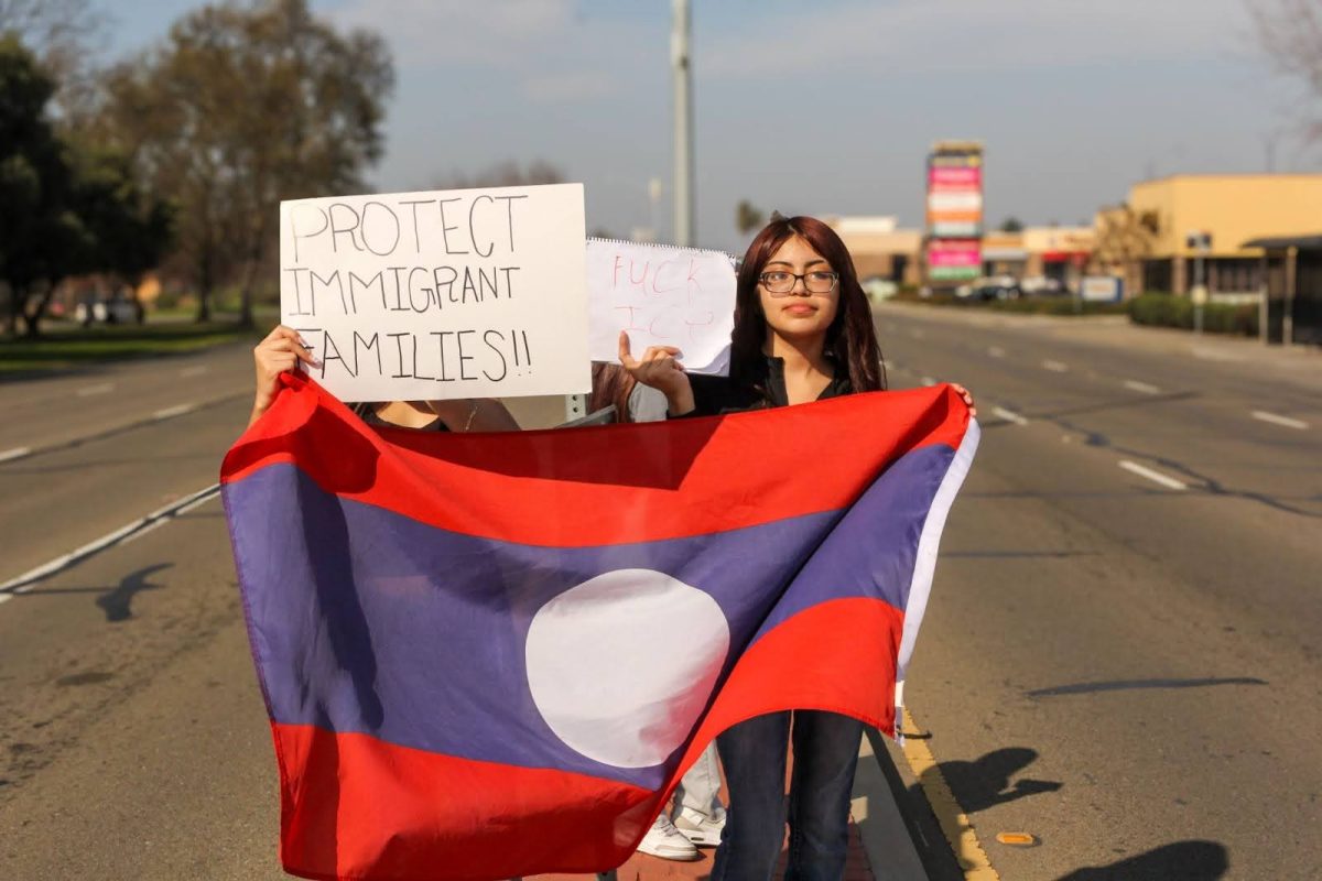 A student holds a “Protect Immigrant Families!!” sign while standing in the roadway with a flag displayed in front.